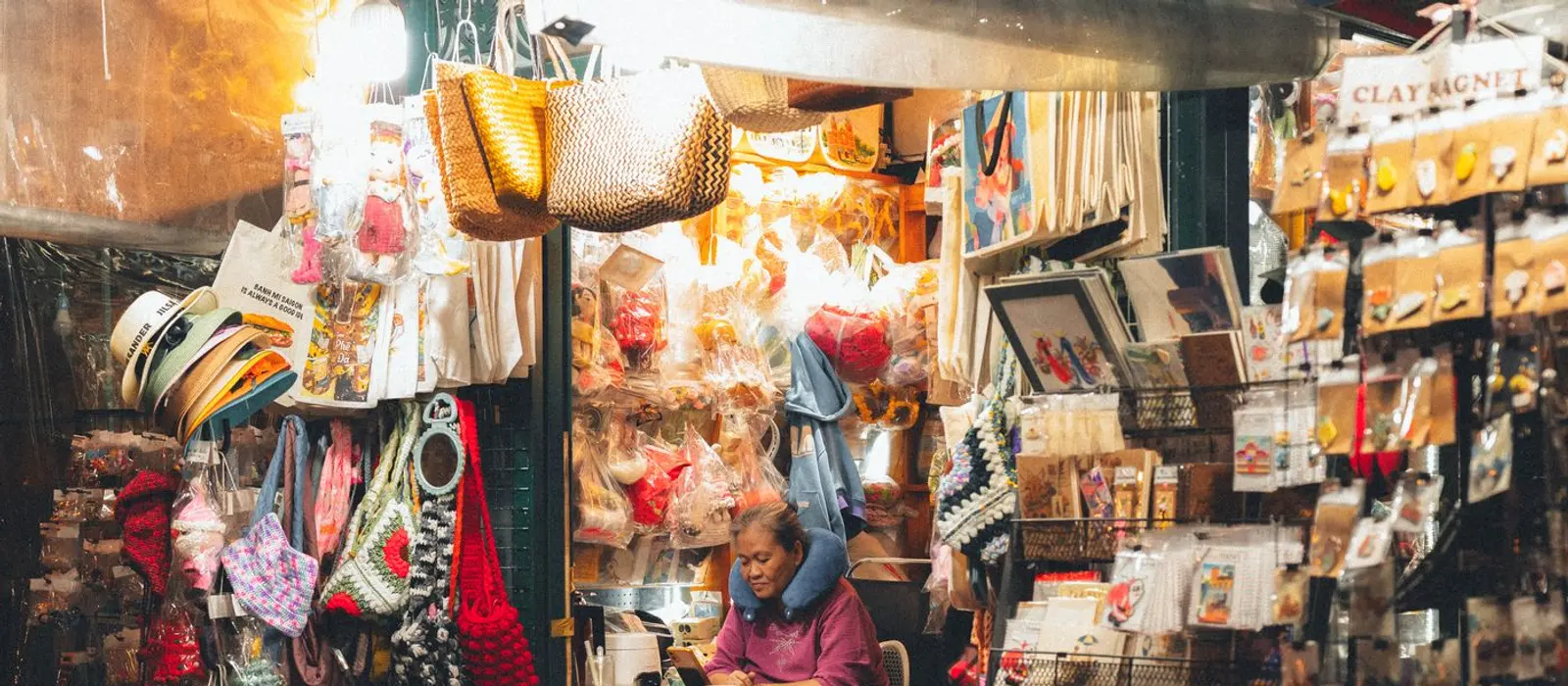 A small market stall packed with handmade goods, lit from within, the stallholder waiting behind the counter.