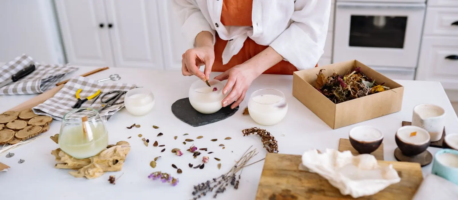 A candle maker's table: poured candles setting, dried flowers, scissors and offcuts spread across the surface.